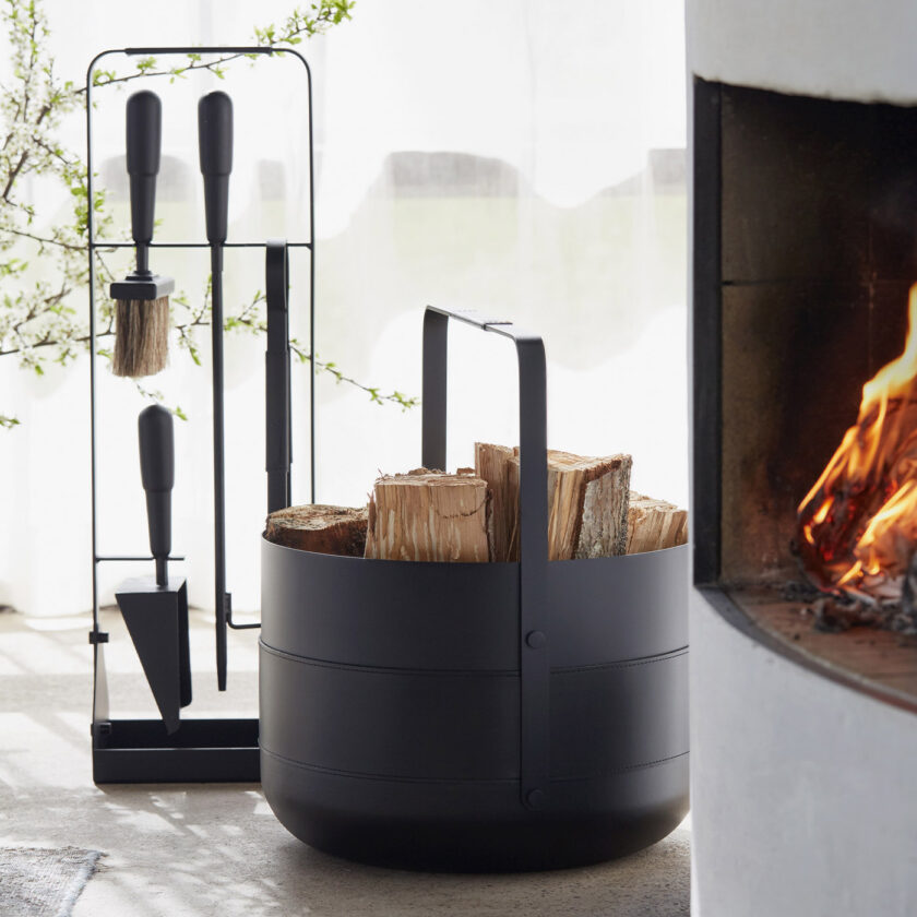 Emma Basket in Noir (charcoal black powder-coated steel, black leather and black details) filled with firewood. It is next to an active fireplace and tools in the same colour.