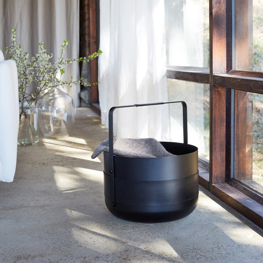 Emma Basket in Noir (charcoal black powder-coated steel, black leather and black details) holding a blanket, by a window.