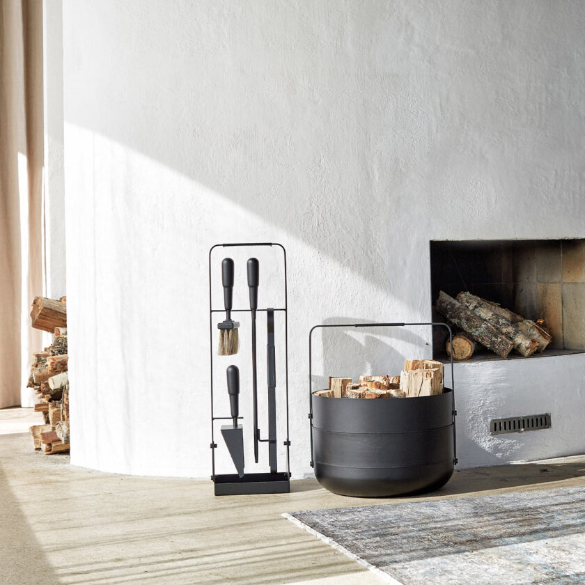 Emma Basket in Noir (charcoal black powder-coated steel, black leather and black details) next to a fireplace and set of tools in the same colour.