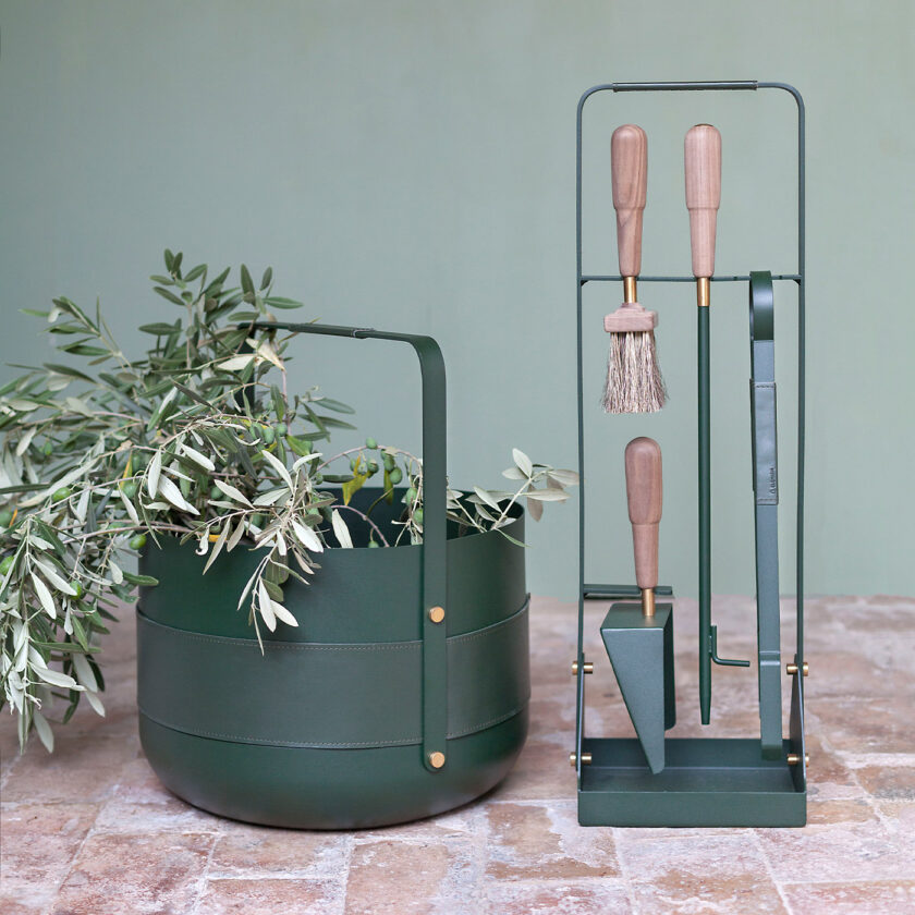 Emma Basket in Forêt (green powder-coated steel, green leather and solid brass details) outdoors with the fireplace tools in the same colour.