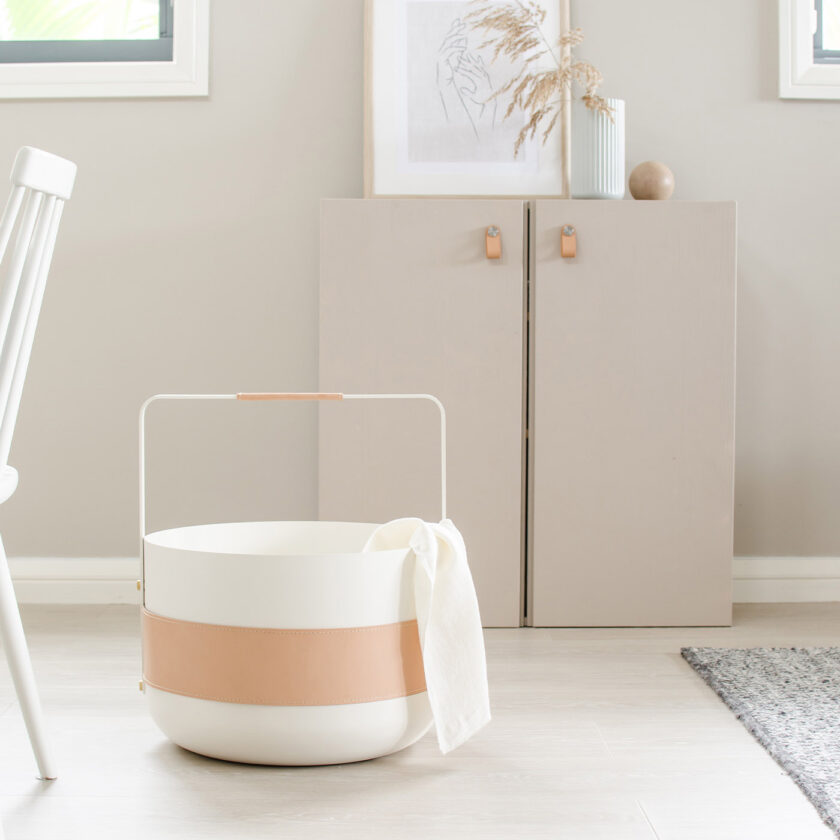 Emma Basket in Blanc (cream white powder-coated steel, beige leather and brass details), holding a white blanket.