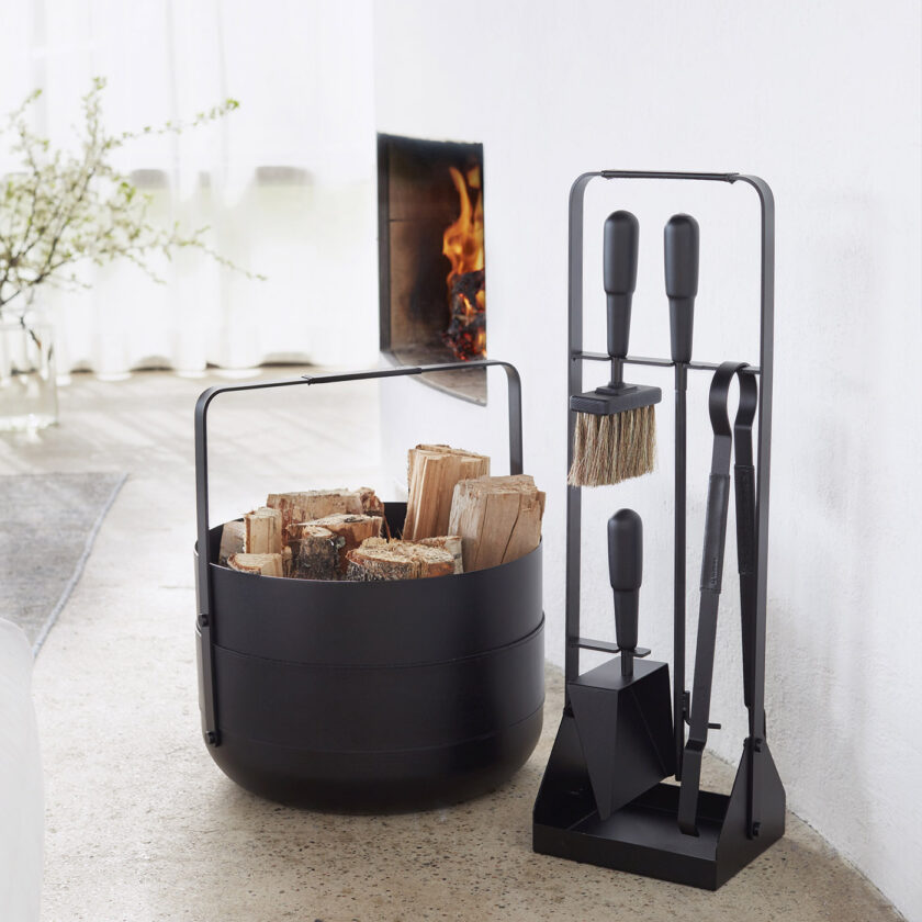 Emma Companion Set in Noir (charcoal black powder-coated steel, “Noir” leather, black stained beech wood handles and black details) next to a basket in the same colour and a fireplace with a lit fire.