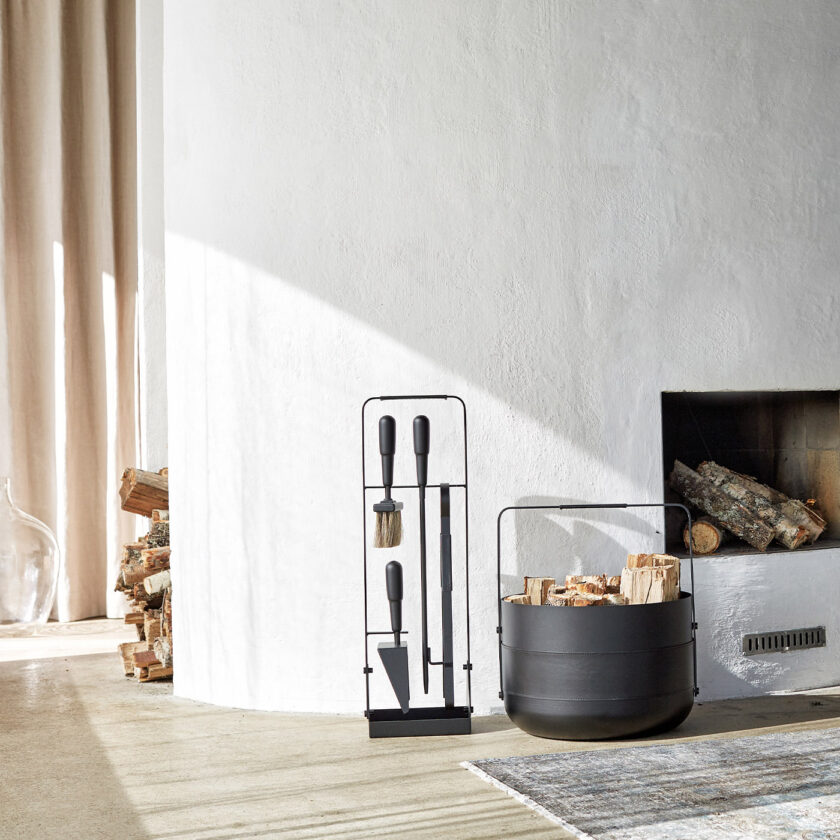 Emma Companion Set in Noir (charcoal black powder-coated steel, “Noir” leather, black stained beech wood handles and black details) next to a basket in the same colour and a fireplace.