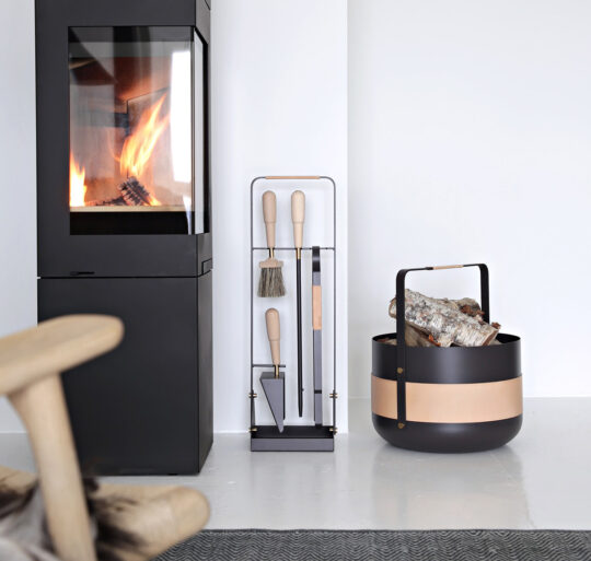 Emma Companion Set in Naturel (dark warm grey powder-coated steel, “Naturel” beige leather, beech wood handles and details in solid brass) next to a basket in the same colour and a fireplace.