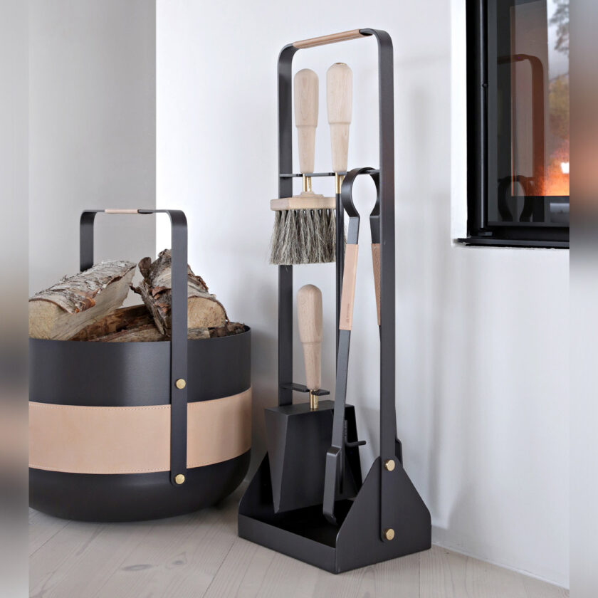 Emma Companion Set in Naturel (dark warm grey powder-coated steel, “Naturel” beige leather, beech wood handles and details in solid brass) next to a basket in the same colour and a fireplace.