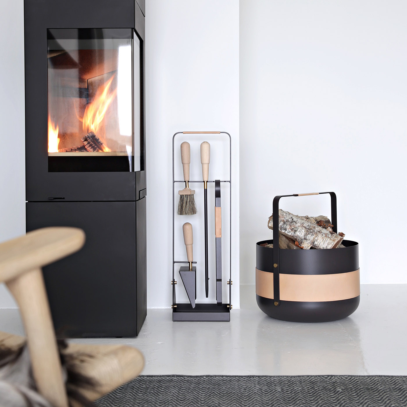 Emma Companion Set in Naturel (dark warm grey powder-coated steel, “Naturel” beige leather, beech wood handles and details in solid brass) next to a basket in the same colour and a fireplace.