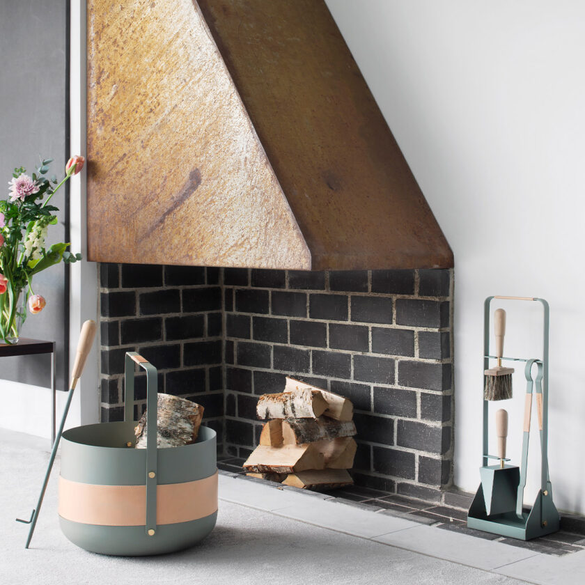 Emma Companion Set in Lichen (light green grey powder-coated steel, “Naturel” beige leather, beech wood handles and details in solid brass) next to a basket in the same colour and a fireplace.