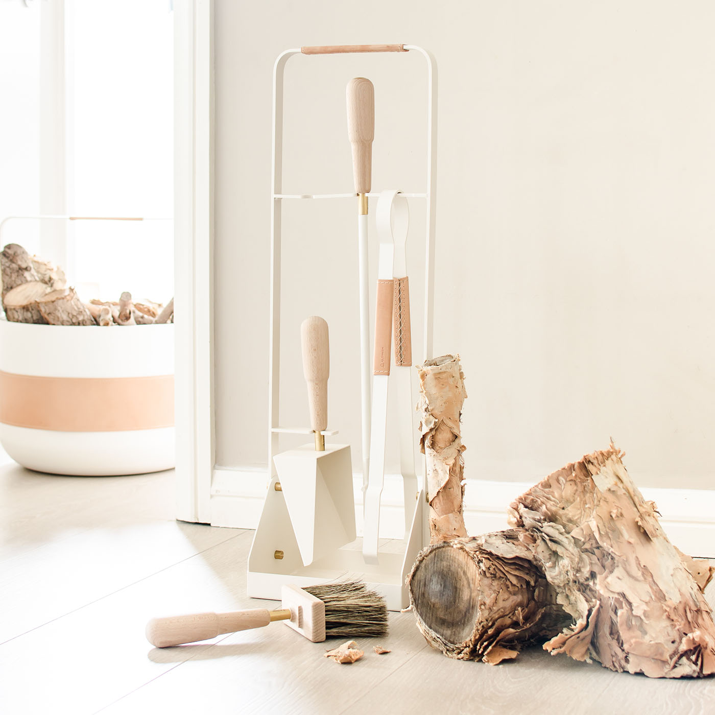 Emma Companion Set in Blanc (cream white powder-coated steel, “Naturel” beige leather, beech wood handles and details in solid brass) next to firewood and a basket in the same colour.