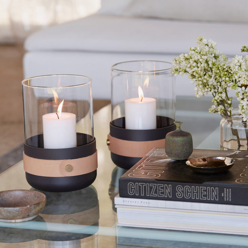Two Emma Lanterns in Naturel (dark warm grey powder-coated steel, “Naturel” beige leather, hand blown glass and brass details) next to books on a glass coffee table close up.