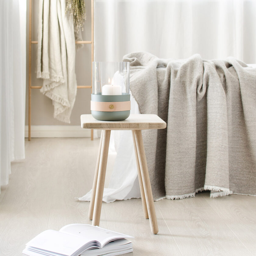 Emma Lantern in Lichen (light green grey powder-coated steel, “Naturel” beige leather, hand blown glass and brass details) on a stool in a bedroom.
