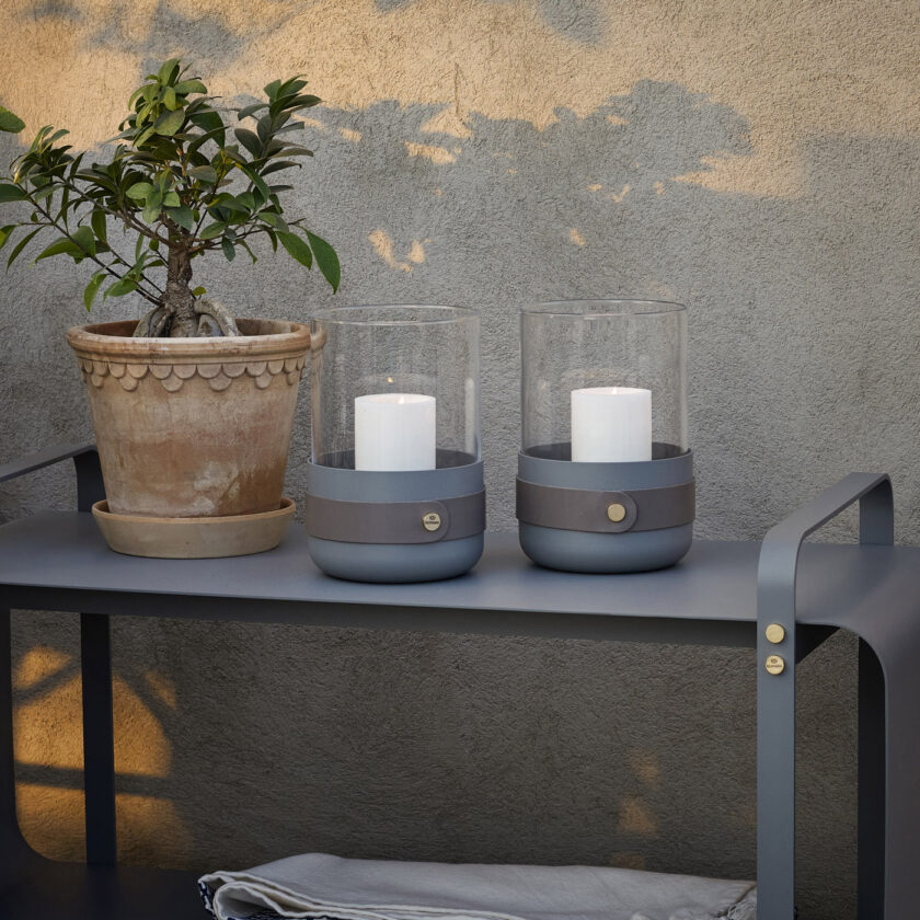 Two Emma Lanterns in Paris (medium grey powder-coated steel, “Ellie” grey leather, hand blown glass and brass details) outside on a bench in the same colour.
