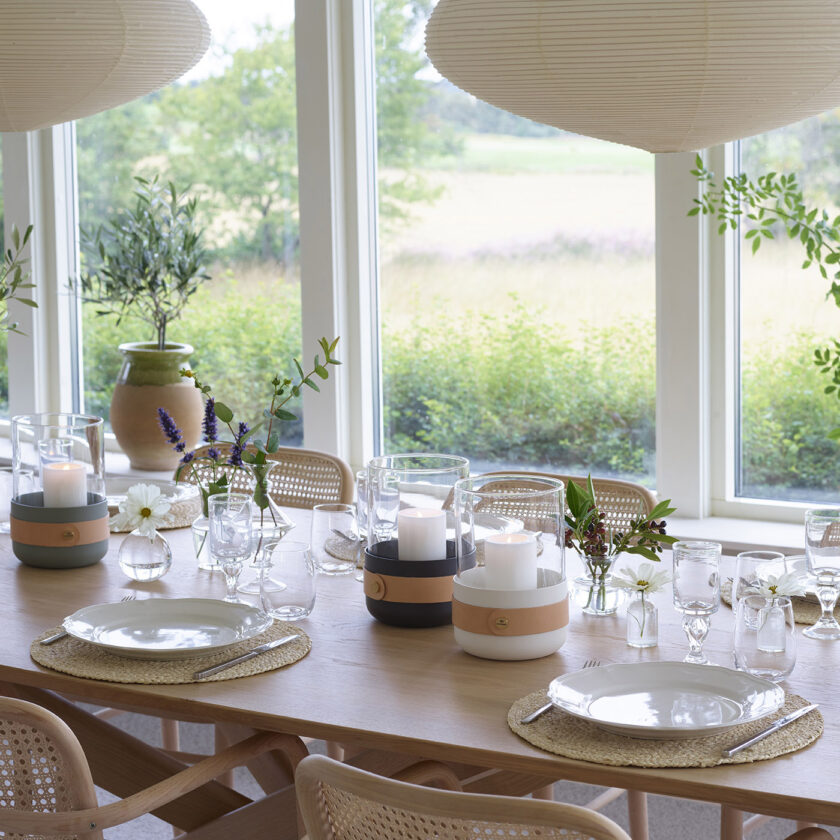 A Blanc Emma Lantern (cream white powder-coated steel, “Naturel” beige leather, hand blown glass and brass details) next to lanterns in other colours on a dinner table.