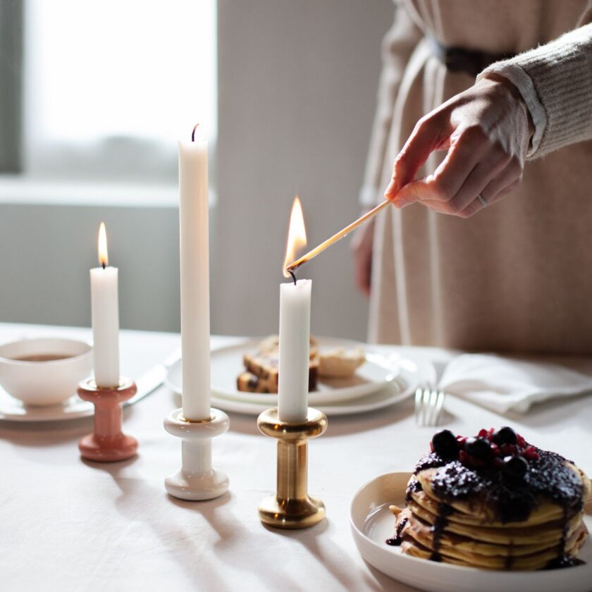 A woman lighting a candle in a Lucie Candle Holder in brass, showing alongside the pink and white versions.
