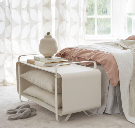 Ninne Bench in white (Blanc) in front of a bed holding pillows inside and books on top.