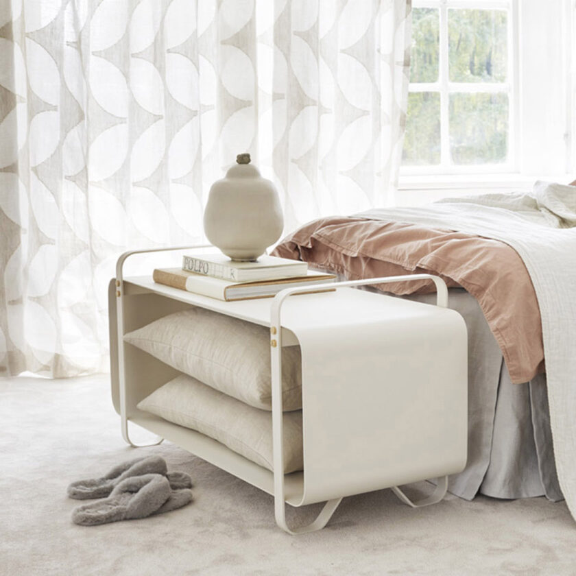 Ninne Bench in white (Blanc) in front of a bed holding pillows inside and books on top.