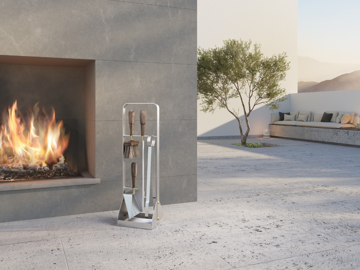 Emma Companion Set in Lumière (combines brushed stainless steel with tool handles in oiled walnut and details in solid brass) outside in front of a lit outdoor fireplace.