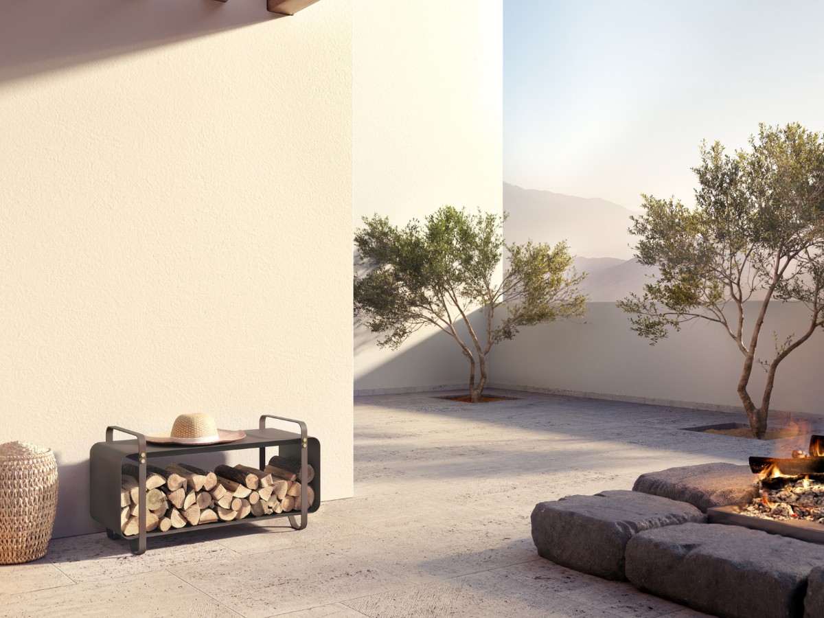 Ninne Bench in Classique (dark warm grey powder-coated galvanised steel with details in solid brass) with wood stacked in it, a hat on top outdoors with a lit outdoor fireplace.