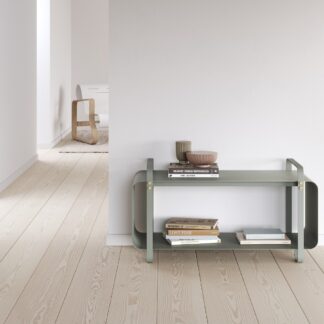 Ninne Bench in Lichen (light green grey powder-coated galvanised steel with details in solid brass) in a white room with books stacked on top and on the inside.