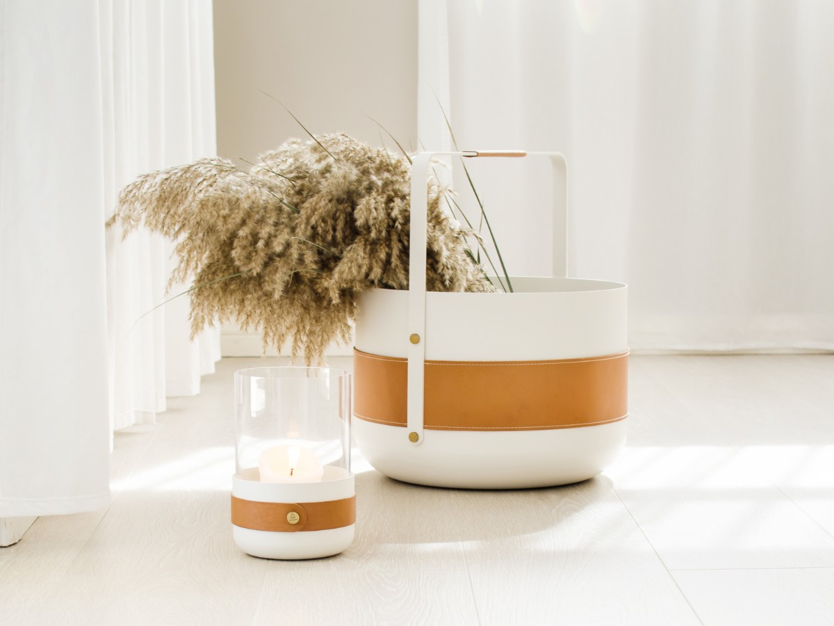 Emma Lantern and Basket in white (cream white powder-coated steel base with hand-stitched “Naturel” leather and details in solid brass). The lantern is lit with a candle and the basket has dried wheat flowers.