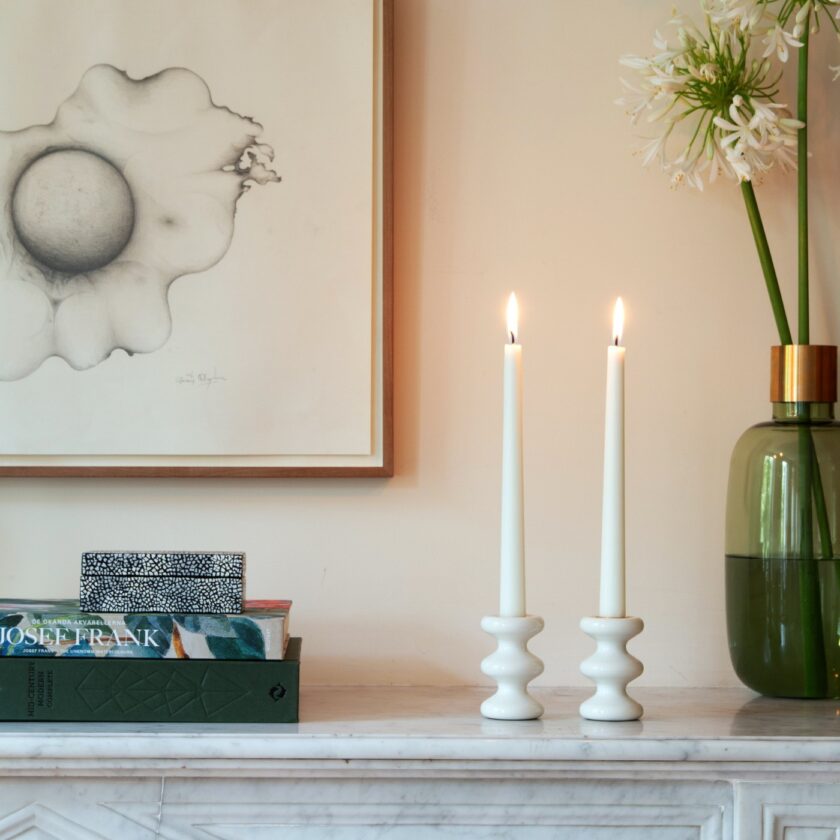 A pair of lit LouLou Candleholders in white (Blanc) on a mantlepiece next to books and flowers.