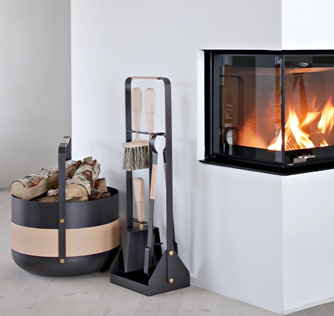 Emma Companion and Basket in Naturel (dark warm grey steel with beige leather and brass details) in front of lit fire.