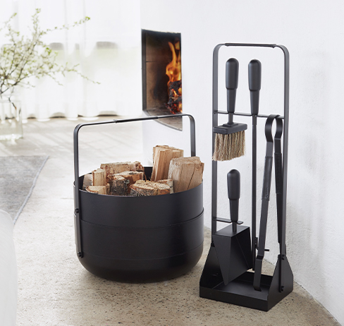 Emma Companion Set and Basket in Noir (charcoal black powder-coated steel, leather and anodised black aluminium details) in front of a lit fire in a white living room.