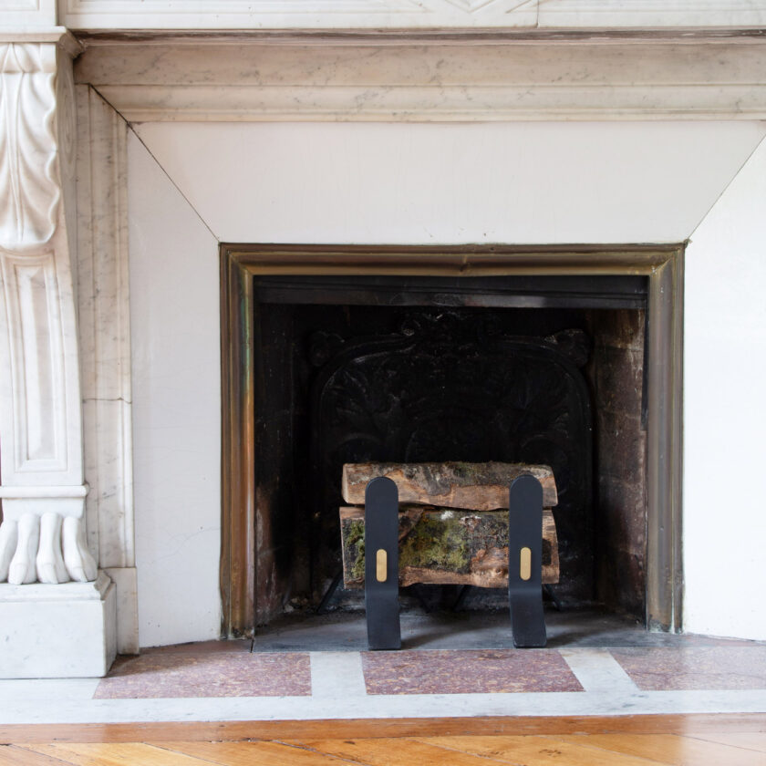 The Noir Cooper Fire Dogs with fire wood on them are placed in an ornate white marble fire place.