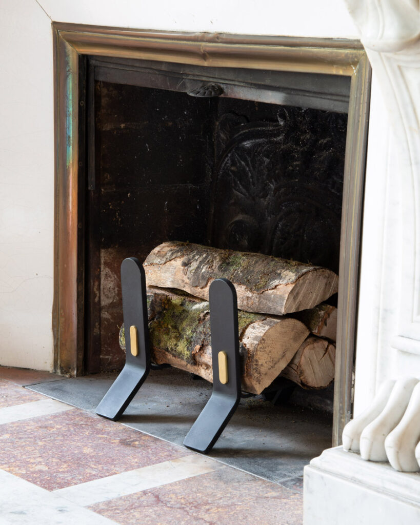 A pair of small Noir Cooper Fire Dogs with wood on them in a fireplace. They have a brass oval shaped detail.