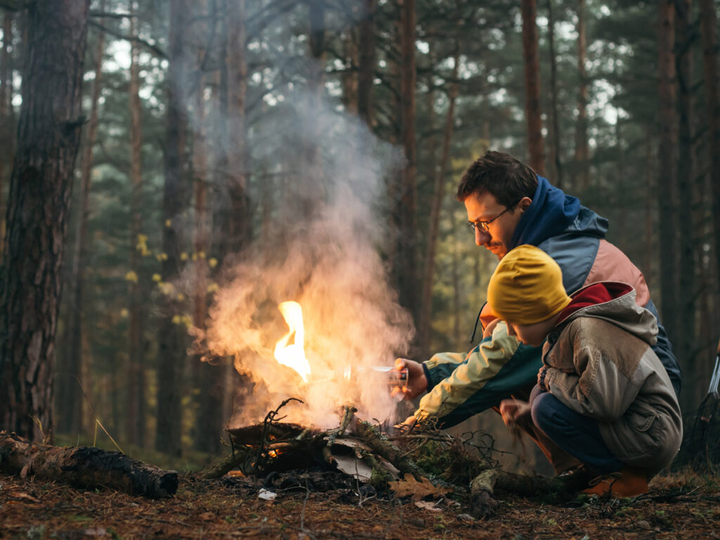 A father and a son in the woods. The father is lighting a camp fire.