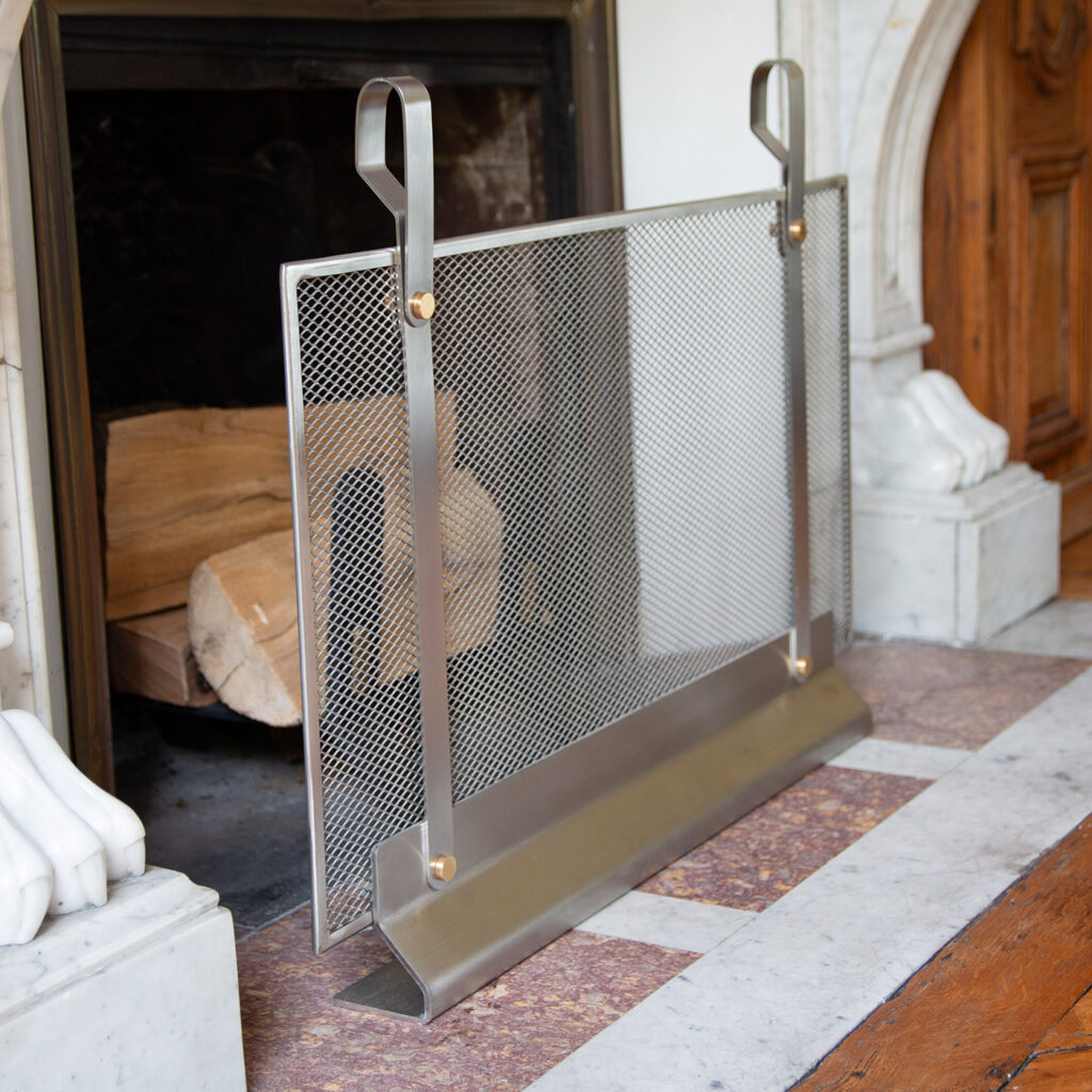 A side view of the Emma Firescreen Small in Lumière in front of a white marble fireplace. Made from stainless steel and solid brass this version can also be used outdoors.