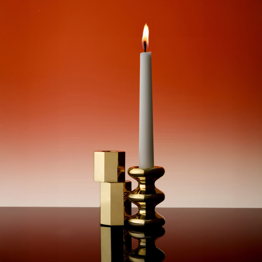 A Luca Candle Holder and a LouLou Candle Holder both in polished brass standing infront of a red background. There is a lit tapered candle in the LouLou Candle Holder.