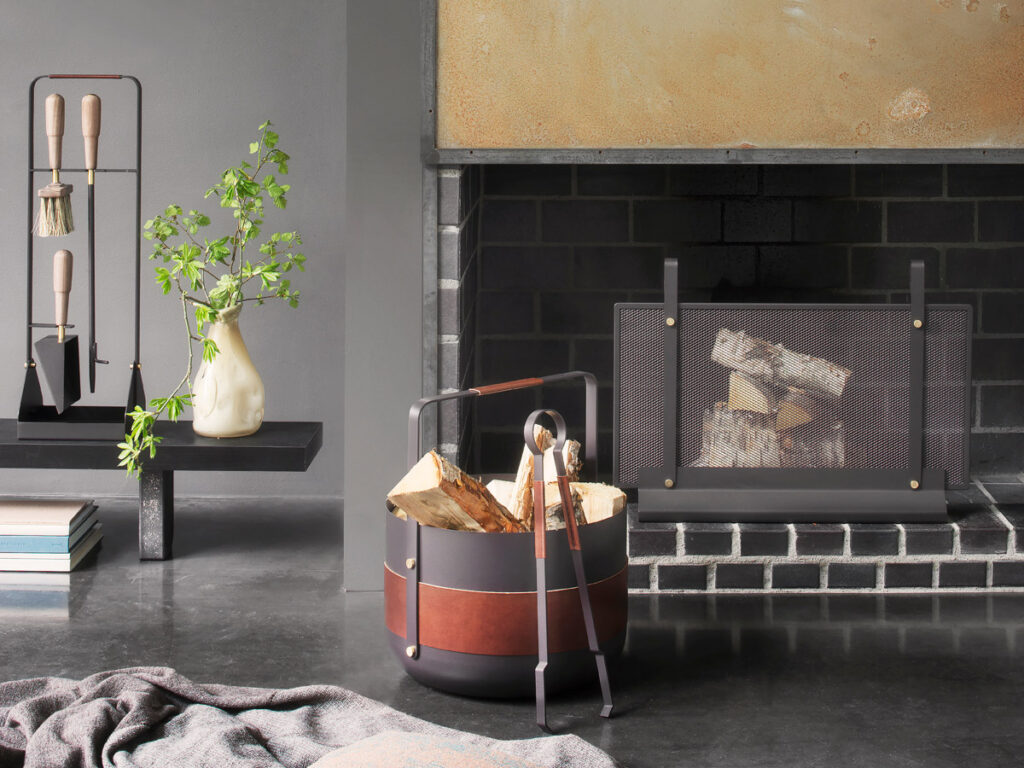 A small Emma Firescreen in Noir with brass in an open fireplace. Next to the fireplace is a Emma Basket and Emma Companion Set in Havane.