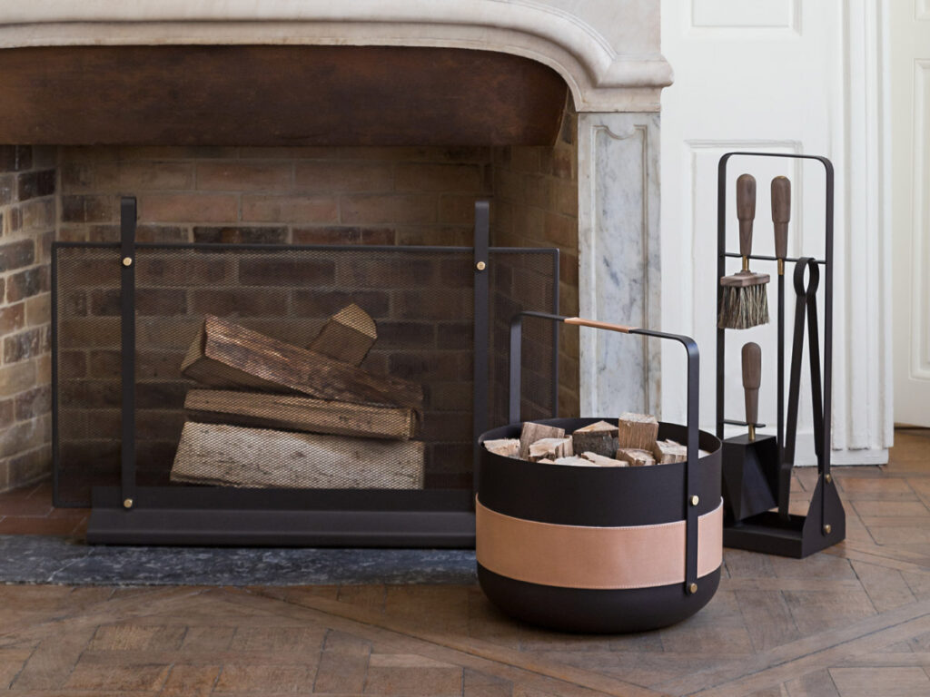 The Emma Firescreen in Classique in front of a marble and brick fireplace. Next to the fireplace is a Emma Basket in Naturel and an Emma Companion Set in Classique.