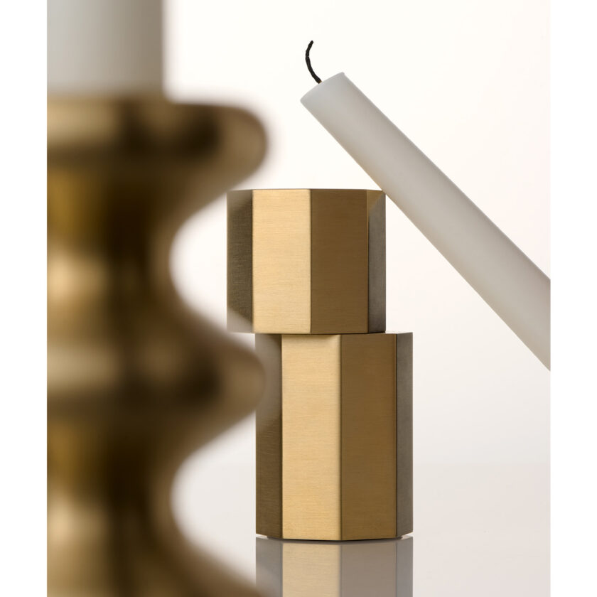 Behind the curves of a Brass LouLou Candle Holder there is an unlit candle leaning against a brushed brass Luca Candle Holder.