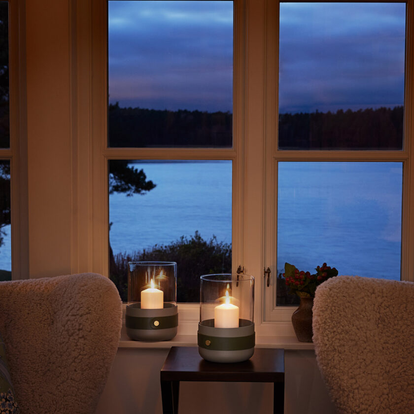 Two Emma Lanterns in Olive with lit candles in them. They are sitting in a window that is looking out to the water at dusk.