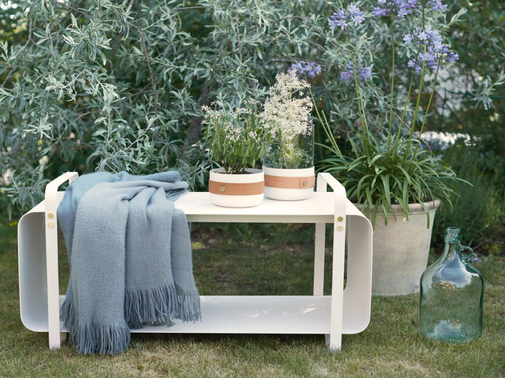 Outdoor Ninne Bench in white colour, shown with two white lanterns containing flowers. and a blue blanket